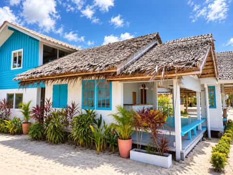 White and turquoise beach house with a thatched terrace, blue benches and tropical plants